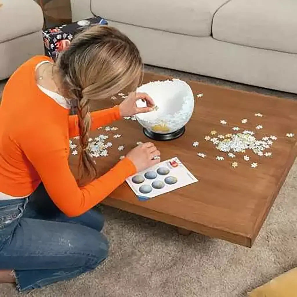Woman in orange sweater kneels by a low table while assembling Ravensburger 3D Puzzle Globe, carefully fitting white and blue interlocking pieces onto the rounded base.