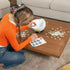 Woman in orange sweater kneels by a low table while assembling Ravensburger 3D Puzzle Globe, carefully fitting white and blue interlocking pieces onto the rounded base.