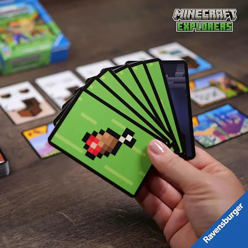 Hand holding pixelated chicken leg food cards from Ravensburger Minecraft Explorers card game, with box and spread-out adventure cards visible on wooden table.