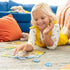 Smiling young girl in a yellow dress lies on a carpet while happily assembling a Ravensburger Peppa Pig puzzle, with blue sofa and orange pillows in background.