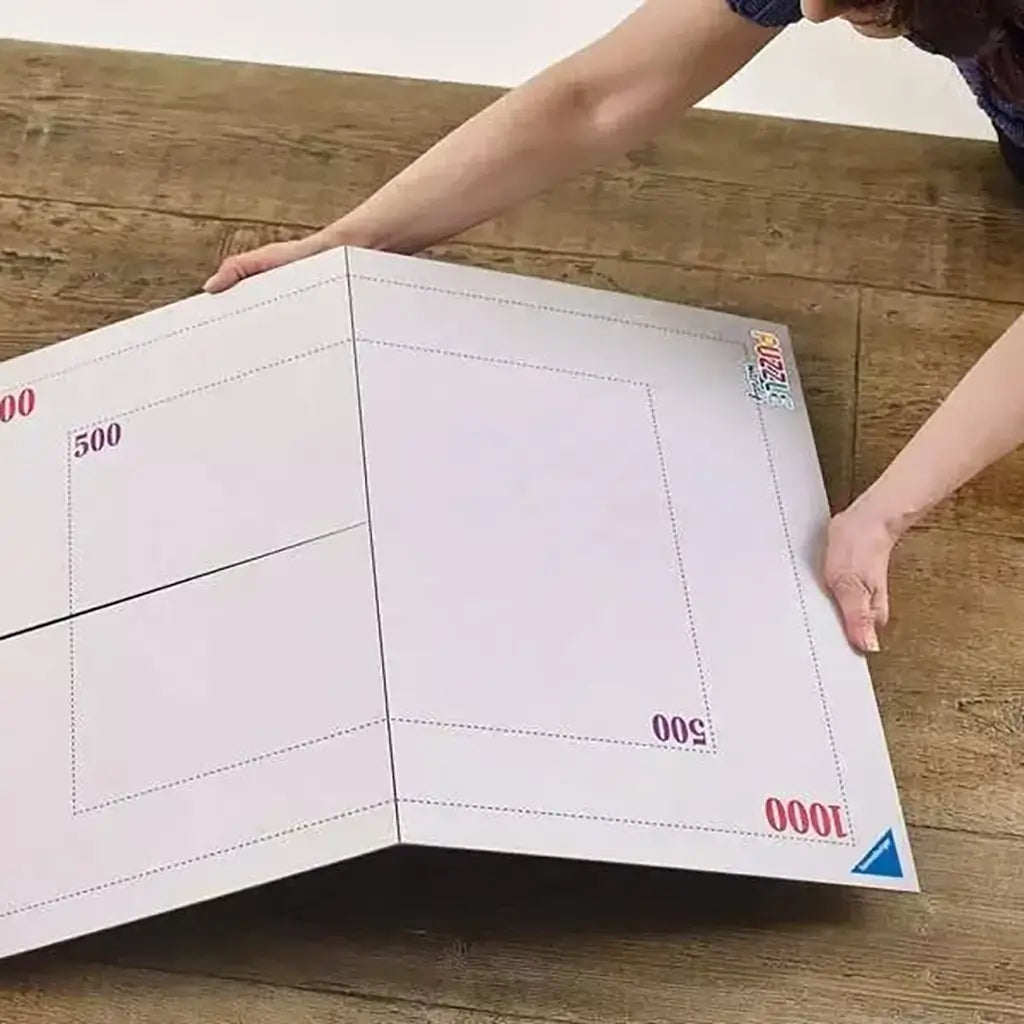 White foldable Ravensburger puzzle board with printed size indicators for 500 and 1000-piece puzzles, shown unfolded on a wooden floor with hands spreading it flat.