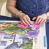 Close-up of hands assembling a jigsaw on the Ravensburger foldaway puzzle board, showing vibrant pink and purple flowers and a stone house against green foliage.