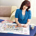 Puzzle board with scattered pieces and a partially built 1000-piece jigsaw, as a woman in a teal cardigan fits pieces together beside a cup of tea on a living room table.