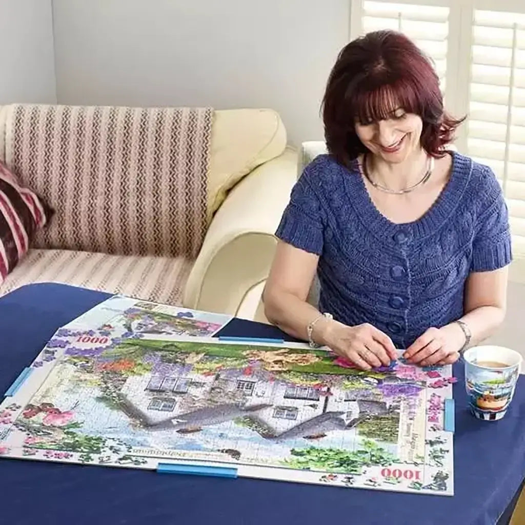 Puzzle board with a colorful 1000-piece jigsaw nearly complete, showing a cottage and flowers, placed on a table as a woman in blue sweater works calmly with a mug beside her.