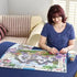 Puzzle board with a colorful 1000-piece jigsaw nearly complete, showing a cottage and flowers, placed on a table as a woman in blue sweater works calmly with a mug beside her.
