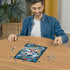 Smiling man in denim shirt assembling Ravensburger Disney Mickey Mouse 300 piece puzzle on round wooden table, colorful blue design nearly completed in center.