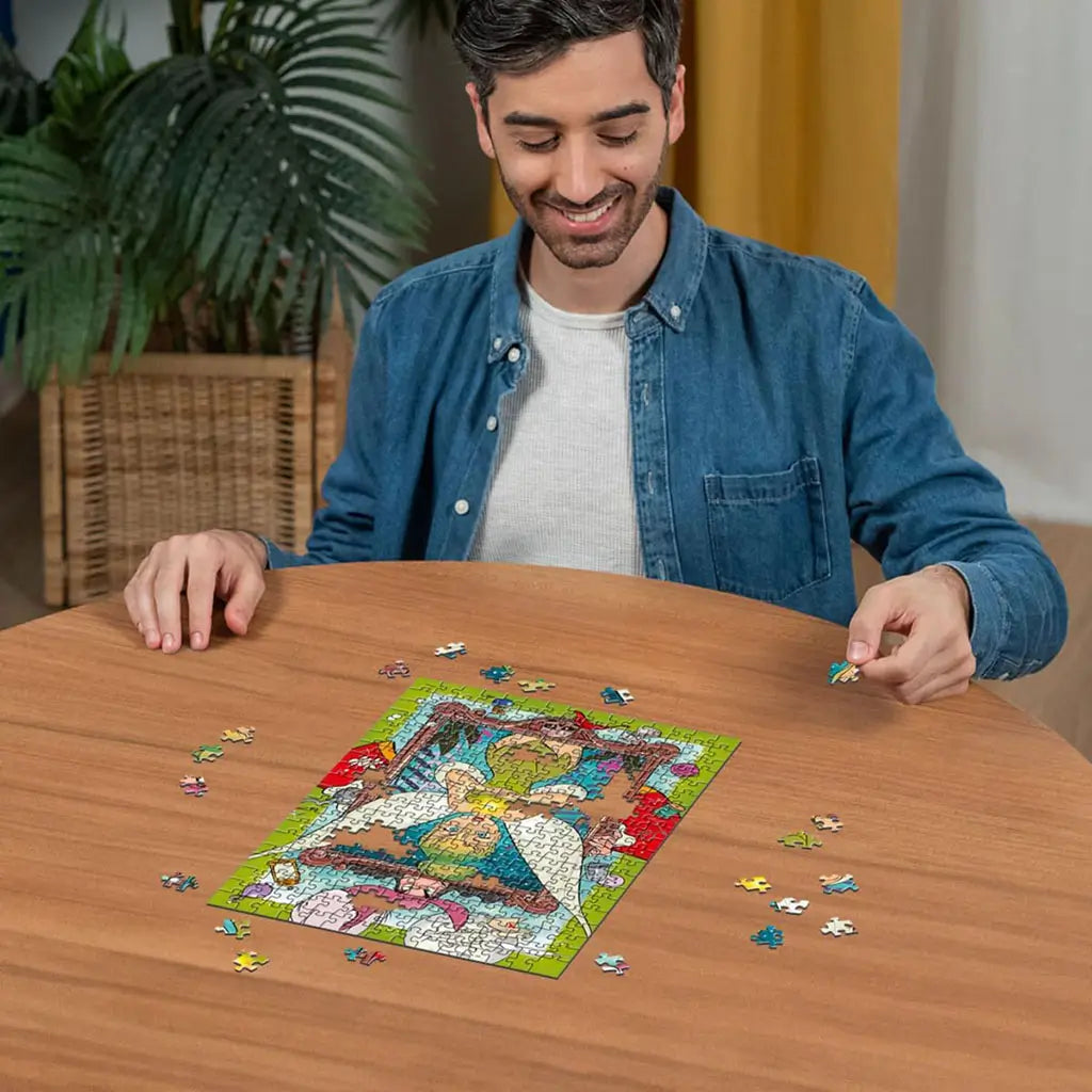 Smiling man in denim shirt sitting at wooden table assembling Ravensburger Disney Tinker Bell 300 piece puzzle, with colorful completed design visible at the center.