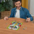 Smiling man in denim shirt sitting at wooden table assembling Ravensburger Disney Tinker Bell 300 piece puzzle, with colorful completed design visible at the center.