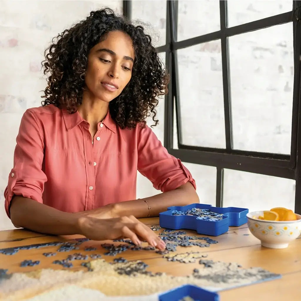 Woman in coral shirt assembling a jigsaw puzzle at wooden table with Ravensburger blue sorting trays and a patterned bowl of orange slices near a large industrial-style window.