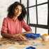 Woman in coral shirt assembling a jigsaw puzzle at wooden table with Ravensburger blue sorting trays and a patterned bowl of orange slices near a large industrial-style window.