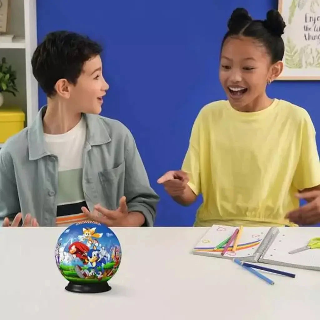 Two excited children sitting at a table with a completed Sonic the Hedgehog 3D puzzle ball between them, surrounded by pencils and school supplies.