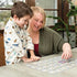 Child in a dinosaur shirt and adult smiling while playing Marvel Spidey and His Amazing Friends matching game together at a light wooden table in a bright living room.