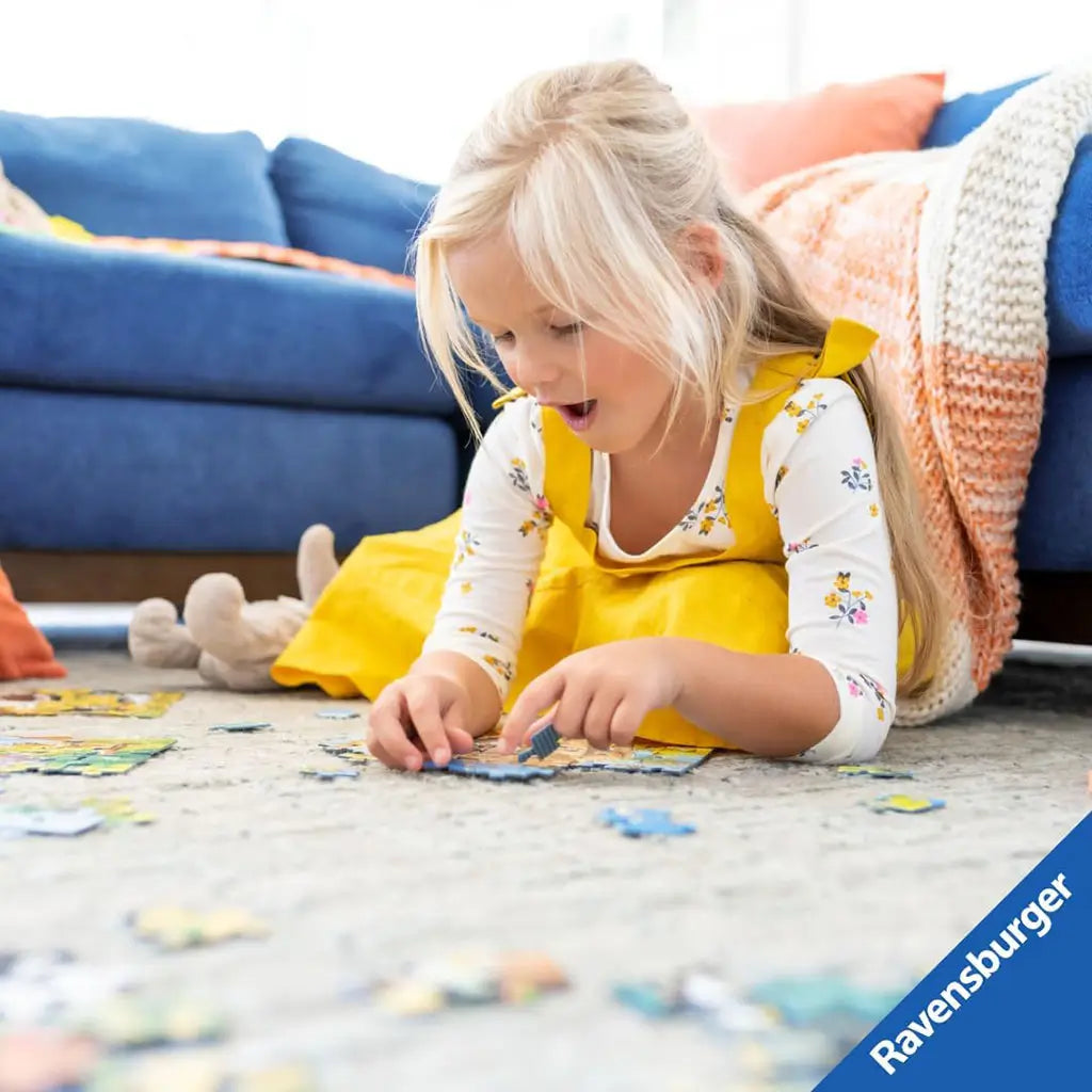 Young blonde girl in a yellow dress lies on a carpet assembling a Ravensburger puzzle, smiling with excitement, while a blue sofa and orange blanket create a cozy living room setting.
