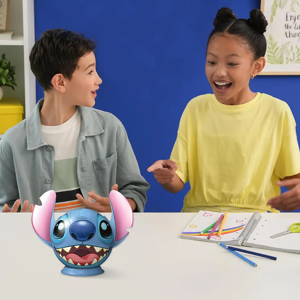 Two excited children at a white table admire a completed Ravensburger Stitch 3D puzzle, surrounded by drawing supplies including colored pencils, notebook, and scissors.
