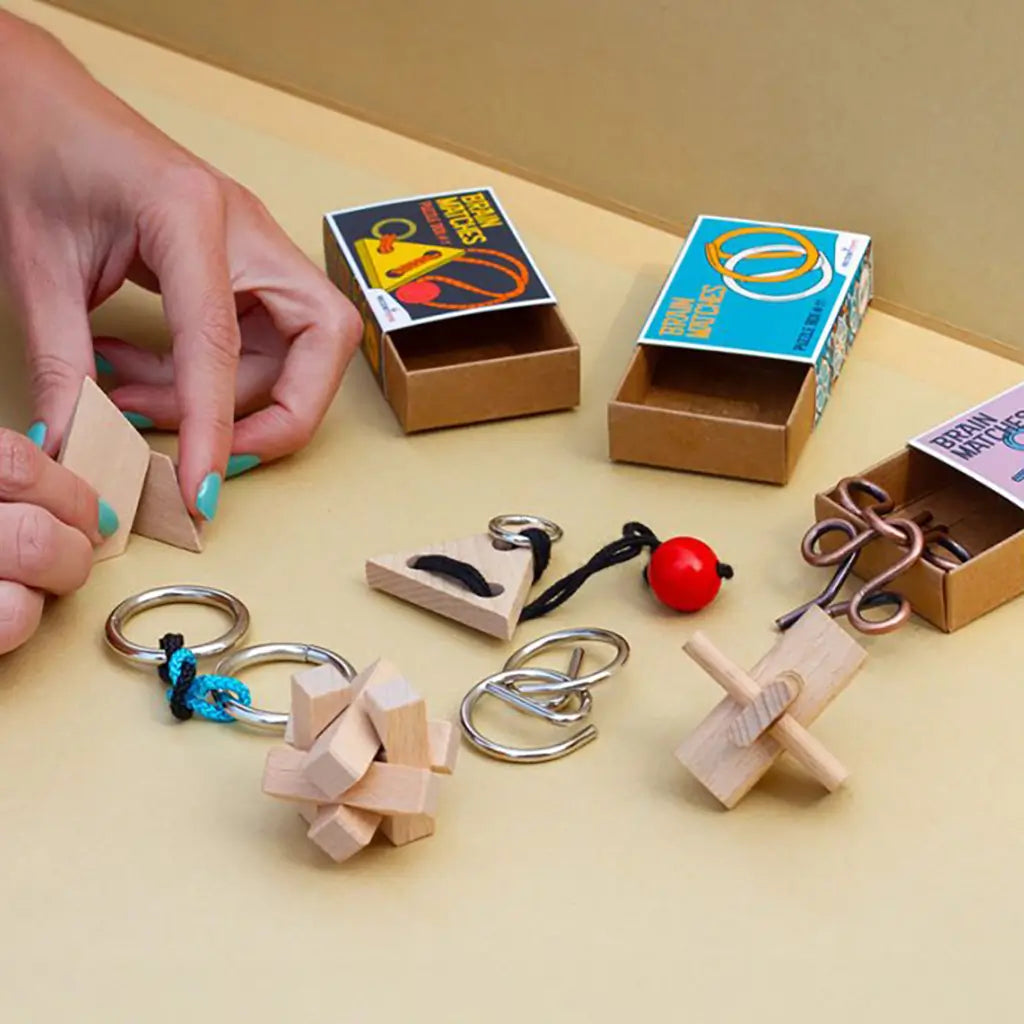Brain teaser puzzles with wood, metal, and rope designs displayed on a table with colorful matchbox-style packaging, showing hands interacting with triangle pieces.