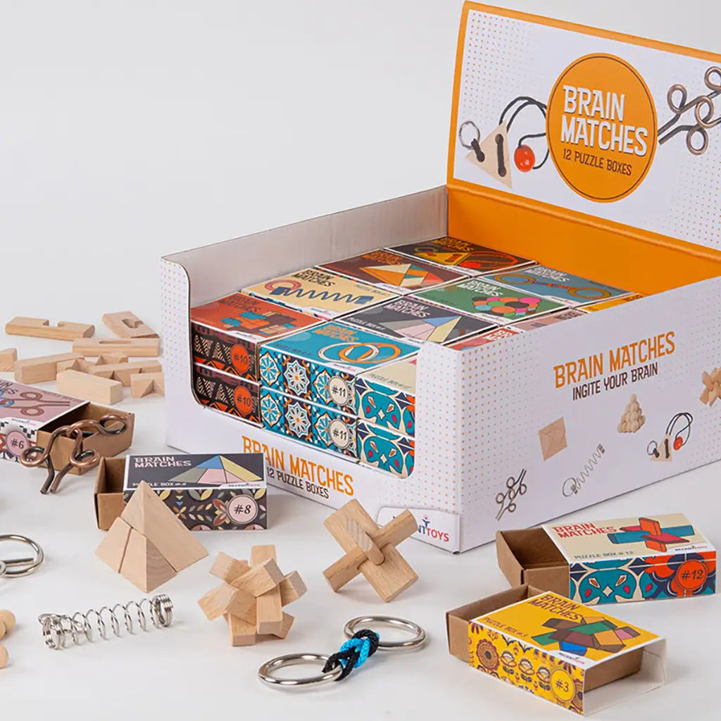 Display box of Brain Matches by Recent Toys containing 12 colorful puzzle boxes with wood and metal challenges spread out in front for interactive play.