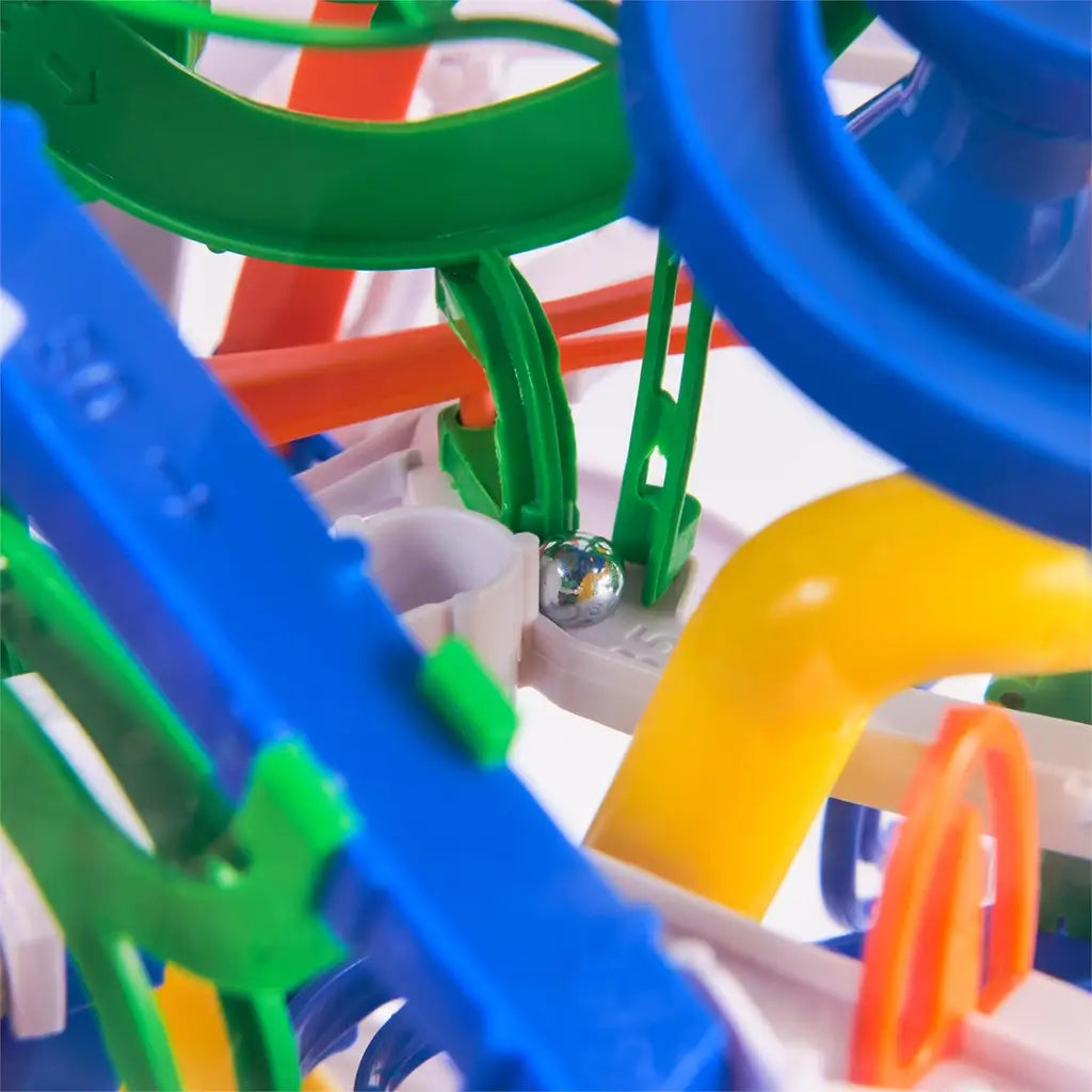 Rubik’s Tracks 3D gravity maze close-up shows green and blue plastic rails with a silver metal ball resting on a white track beside a curved yellow tube inside the clear sphere.