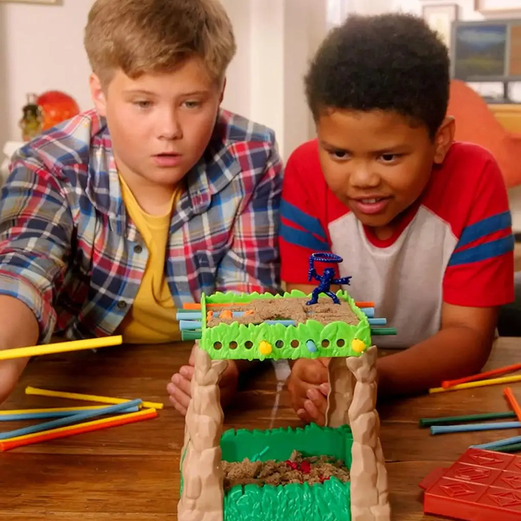 Two boys concentrate while playing Sink N’ Sand game, pulling colorful sticks from jungle platform as explorer figures balance on sand above.