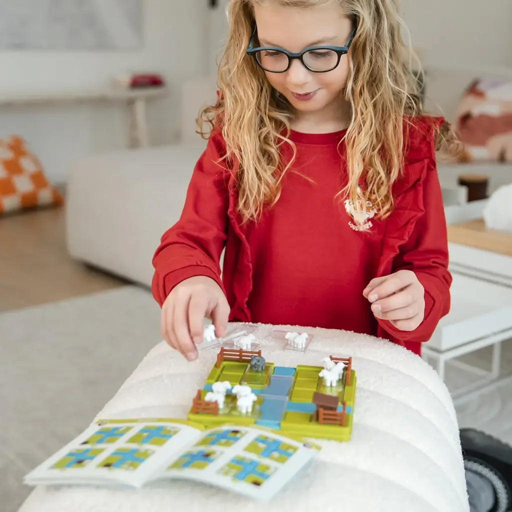 Young girl wearing glasses and a red sweater playing the Smart Games Counting Sheep puzzle on a cushion, using white sheep figurines and following the challenge booklet.