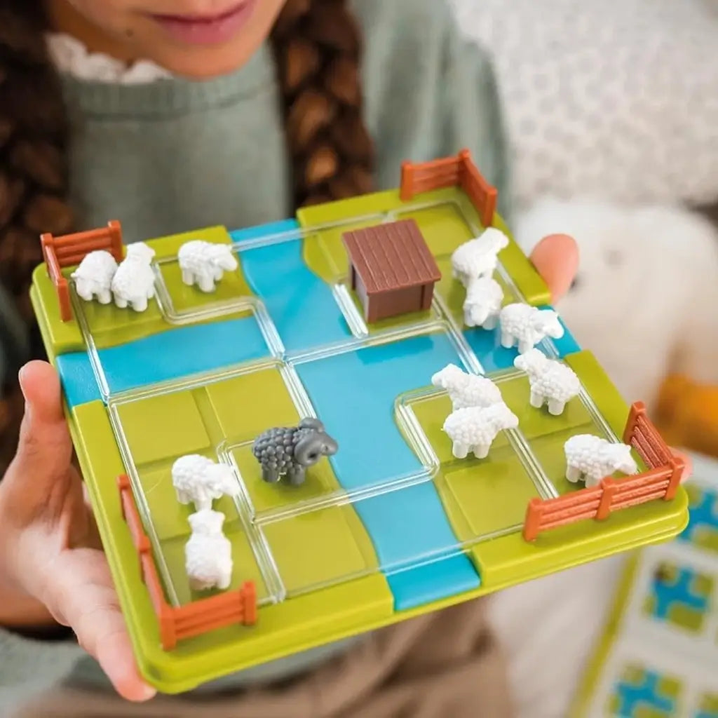 Educational puzzle game board with green base, blue paths, and small white sheep figures, shown in a child’s hands while playing at home on a soft surface.