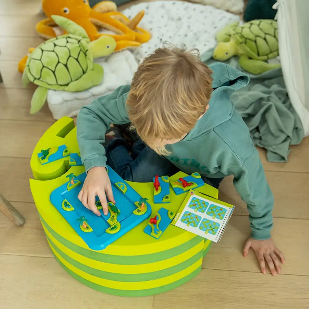 Young boy in green hoodie plays Smart Games Dinosaurs Mystic Islands brainteaser on a striped green table, moving dinosaur pieces while plush turtles and an octopus sit nearby.