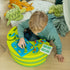 Young boy in green hoodie plays Smart Games Dinosaurs Mystic Islands brainteaser on a striped green table, moving dinosaur pieces while plush turtles and an octopus sit nearby.