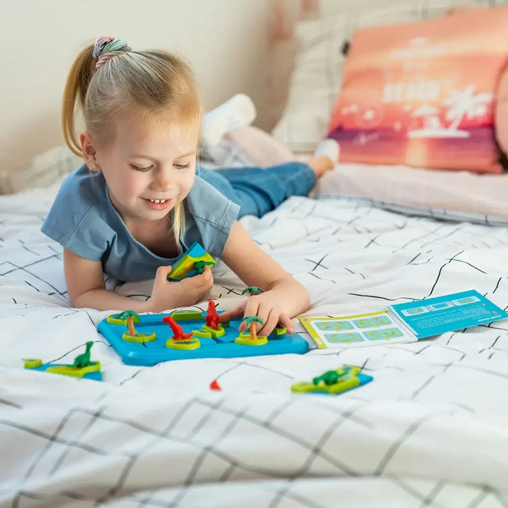 Smiling girl lies on a bed, playing the Smart Games Dinosaurs Mystic Islands brainteaser, holding a puzzle tile while arranging dinosaur figures on the colorful game board.