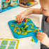 Child plays with the Smart Games Dinosaurs Mystic Islands puzzle, arranging colorful green and red dinosaur figures on blue and green island pieces at a white table.