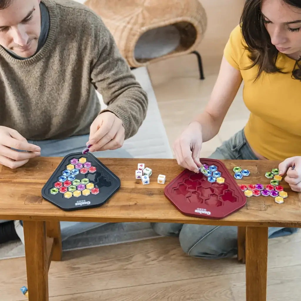 Board game Genius Gems by Smart Games shown on a wooden table with two players placing colorful hexagonal jewel pieces on red and black plastic trays.