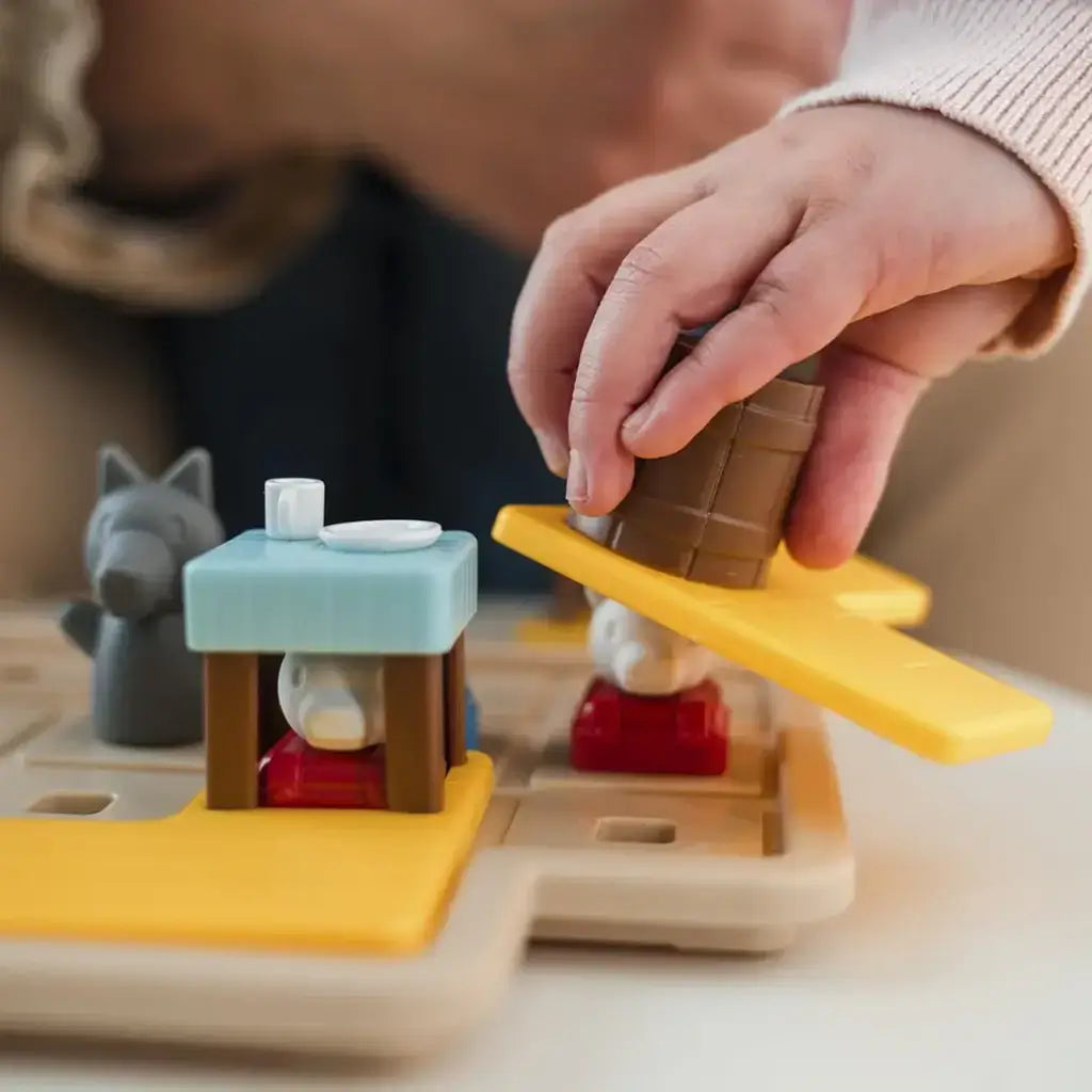 Close-up of child’s hand placing a brown barrel piece on the Smart Games Hide & Goat Seek puzzle, hiding a small goat figure beneath a blue table as part of the preschool brain teaser.