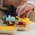 Close-up of child’s hand placing a brown barrel piece on the Smart Games Hide & Goat Seek puzzle, hiding a small goat figure beneath a blue table as part of the preschool brain teaser.