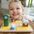 Smiling young girl leaning close to Smart Games Hide & Goat Seek puzzle on wicker table, showing green clock, blue table, and goat figurine as she enjoys interactive playtime.