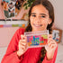 Smiling girl in red knit sweater with braided hair holding a clear Smart Games IQ Bubbles case filled with colorful pieces, seated indoors against a decorated wall background.