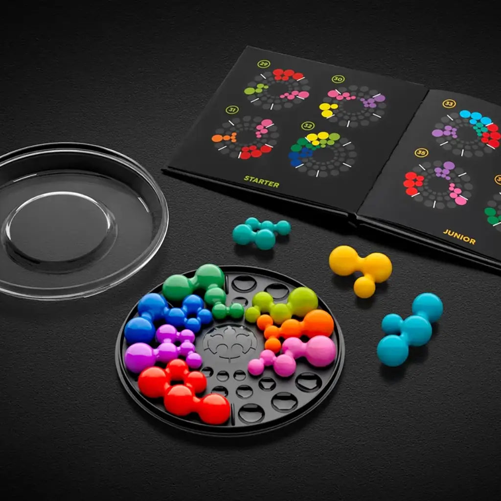 Open SmartGames IQ Circle set with round black tray, multicolored ball-cluster puzzle pieces, clear lid, and illustrated booklet showing starter challenges.