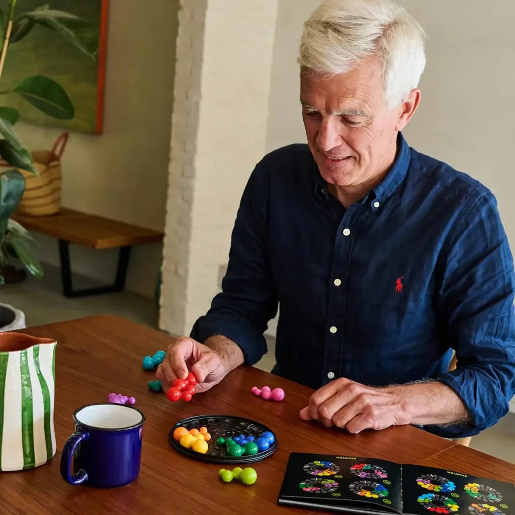 An older man at a wooden table enjoys arranging red ball-shaped pieces in the SmartGames IQ Circle puzzle tray, with other colorful clusters around him.