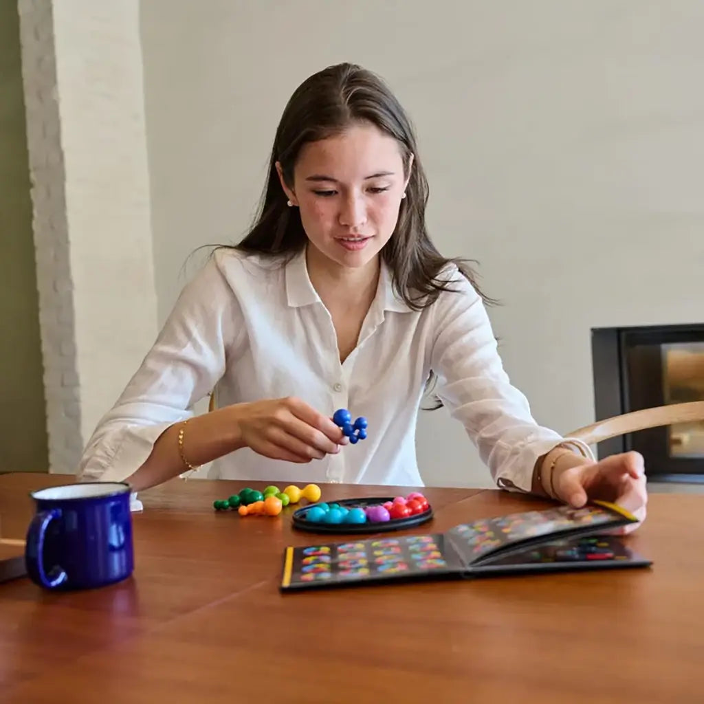 A young woman in a white blouse studies the challenge booklet while holding a blue ball-cluster piece, preparing to fit it into the SmartGames IQ Circle puzzle.