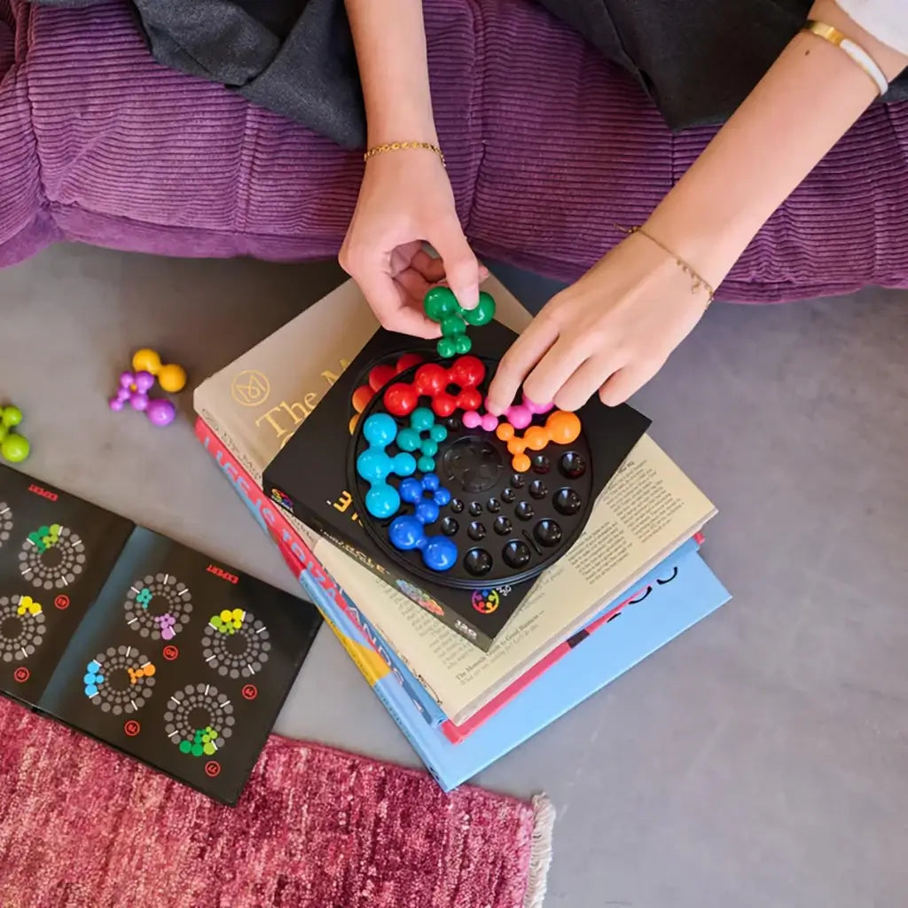 A person sits on a couch stacking SmartGames IQ Circle puzzle pieces on books, solving the colorful ball-cluster logic game with the open booklet nearby.