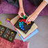 A person sits on a couch stacking SmartGames IQ Circle puzzle pieces on books, solving the colorful ball-cluster logic game with the open booklet nearby.