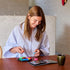 Woman seated at a wooden table, smiling as she fits colorful SmartGames IQ Hexagon pieces onto the puzzle board, with open challenge booklet beside her.