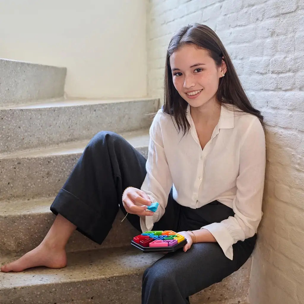 Young woman sitting on stairs in casual clothes, smiling while playing with the colorful SmartGames IQ Hexagon puzzle, holding a bright blue piece above the board.