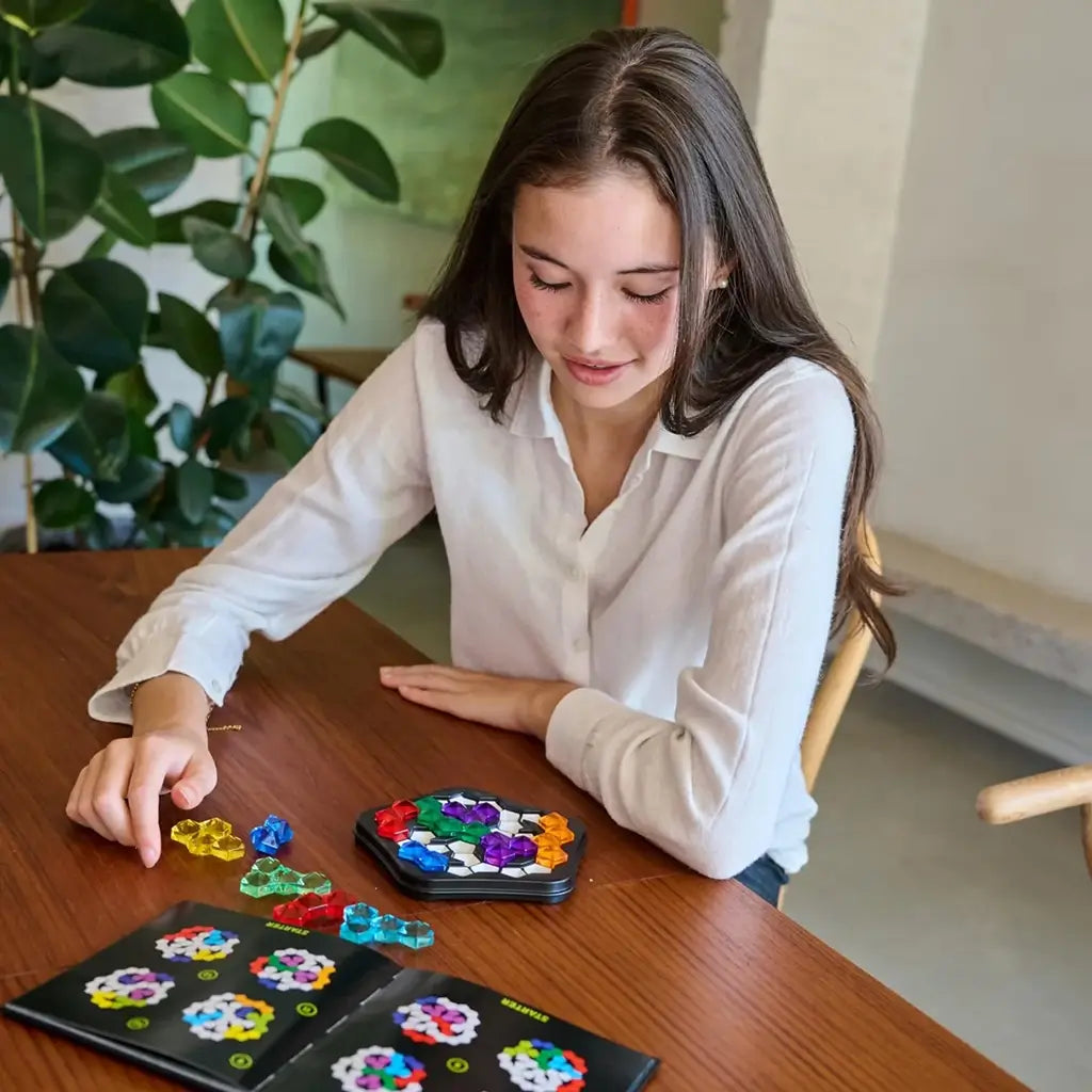 A young woman sits at a wooden table concentrating on placing colorful plastic puzzle pieces into the black pentagon-shaped SmartGames IQ Pentagon logic game