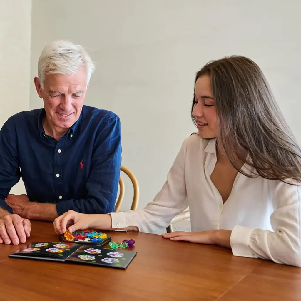 An older man and young woman smile while sitting at a wooden table, working together on the SmartGames IQ Pentagon puzzle with colorful pieces and booklet.