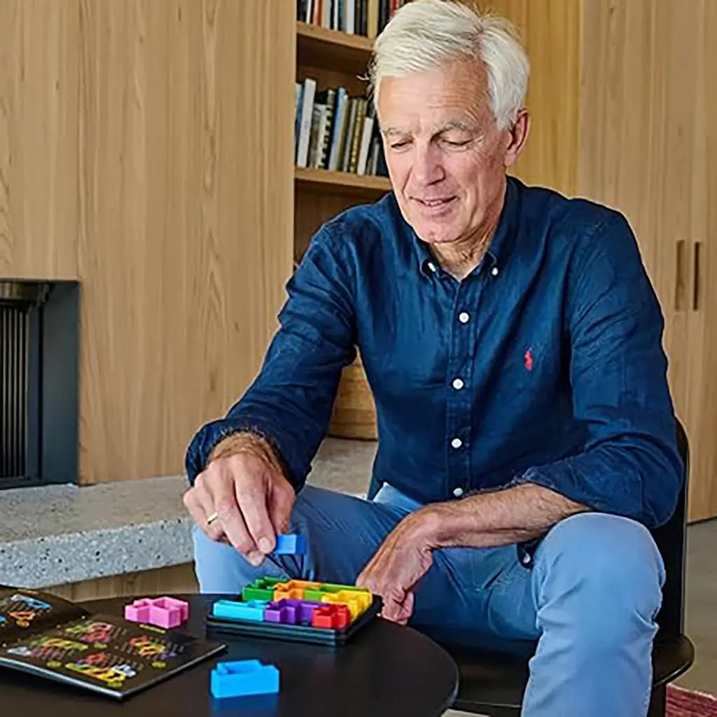 An older man in a navy shirt smiles while placing a blue block into the SmartGames IQ Square puzzle tray, with the open challenge booklet nearby.