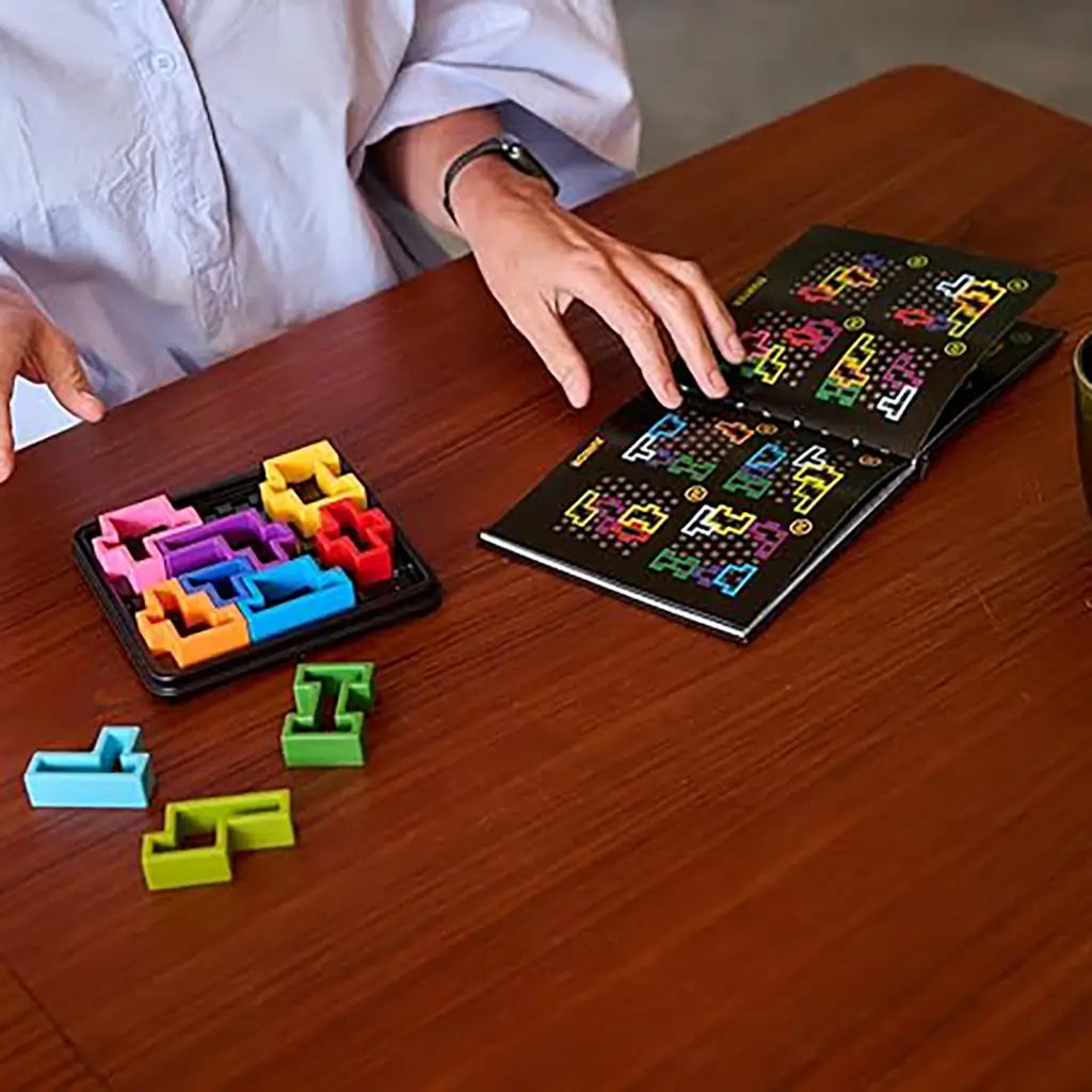 A person at a wooden table plays SmartGames IQ Square puzzle, arranging colorful block pieces while checking the illustrated challenge booklet for solutions.