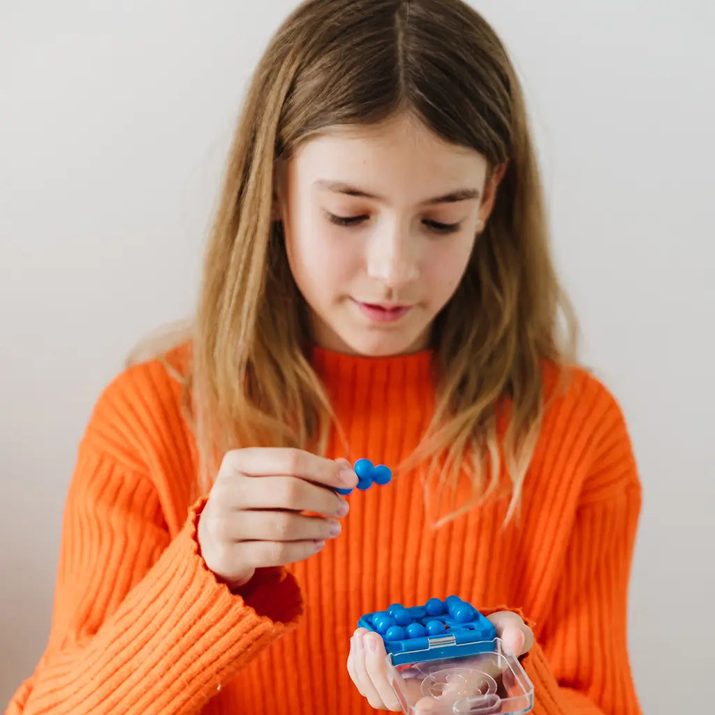 Young girl in an orange sweater holding a blue SmartGames IQ Mini puzzle, carefully fitting bead-shaped pieces into the small transparent plastic case.