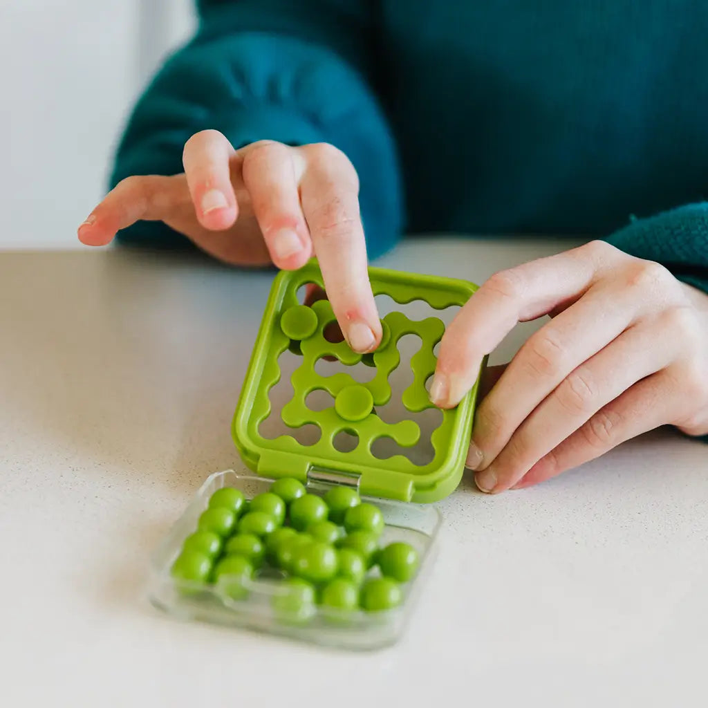 Green plastic SmartGames IQ Mini brainteaser with round puzzle pieces being placed by hand on a smooth table, showcasing its tactile logic challenge design.