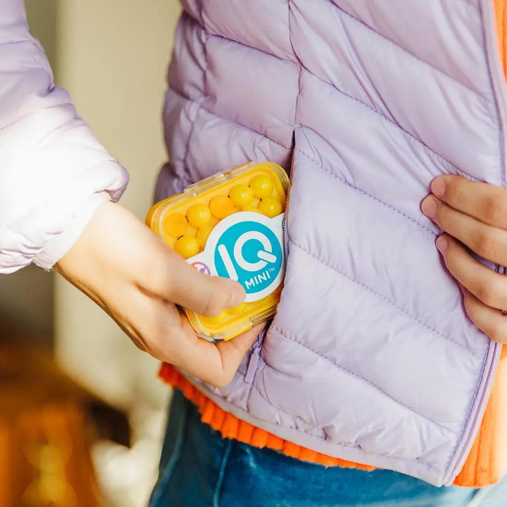 Yellow SmartGames IQ Mini brain puzzle with round beads shown slipping into a lavender jacket pocket, emphasizing its compact and travel-friendly size.