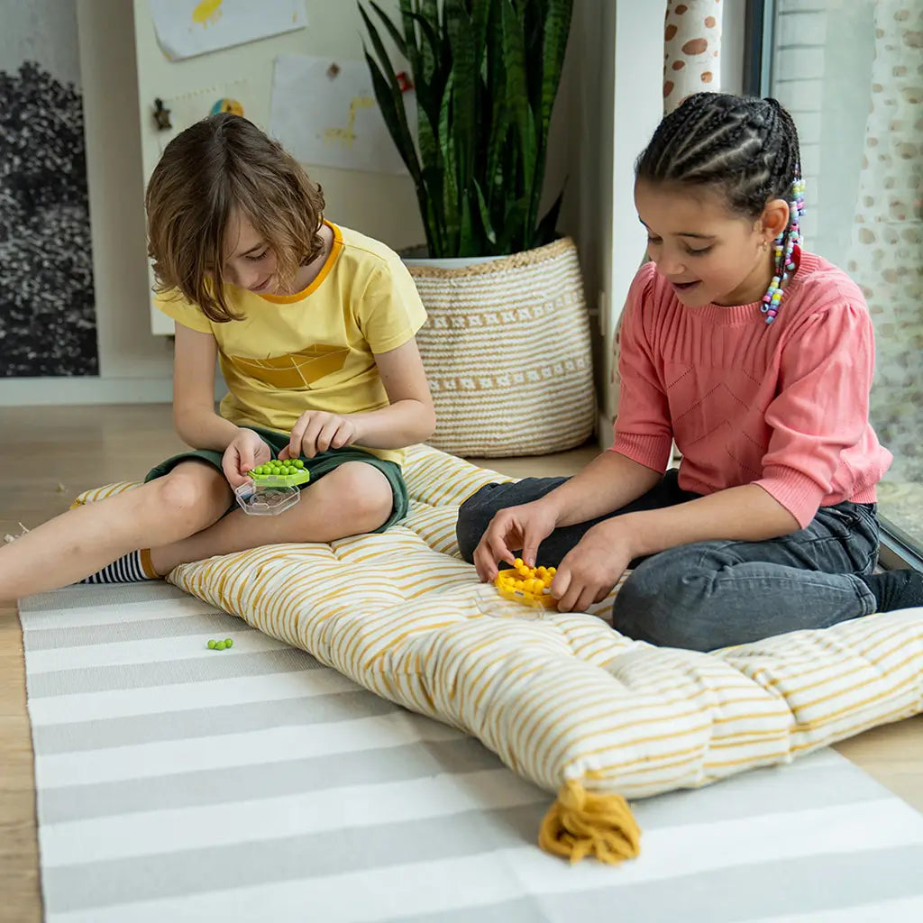 Two children sitting on a striped cushion by a window, playing Smart Games IQ Mini Hexpert puzzles in green and yellow, focusing on fitting the colorful spheres.