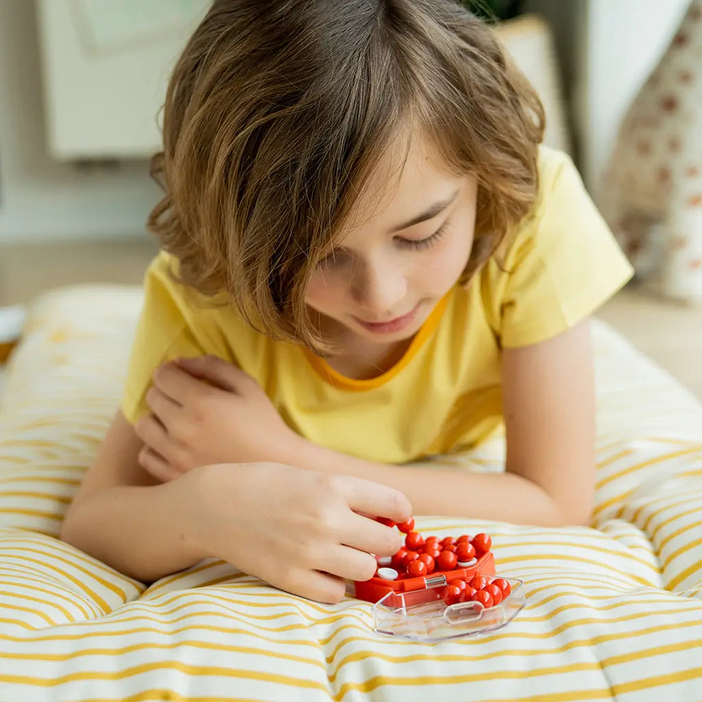 Young child in a yellow t-shirt lying on a striped cushion while concentrating on the red Smart Games IQ Mini Hexpert puzzle, arranging sphere pieces into the case.