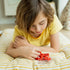 Young child in a yellow t-shirt lying on a striped cushion while concentrating on the red Smart Games IQ Mini Hexpert puzzle, arranging sphere pieces into the case.